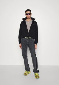 Svart kort jakke med høyt krage, grå t-skjorte, grå jeans med neonskjerf, og flerfargede joggesko. Solbriller fullfører antrekket.