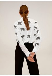 Chemise blanche à manches longues avec un imprimé léopard noir répété, coupe ajustée, tissu lisse et poignets boutonnés. Associée à un pantalon noir.