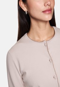 Helle rosa Button-Down-Cardigan mit einem runden Hals, strukturierter Garnverzierung und vier Knöpfen; Model trägt silberne Creolen.