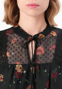 Blusa nera floreale con colletto rotondo, tessuto trasparente a pois, motivi floreali in tonalità di rosso e arancione, e dettagli in pizzo sulle spalle.