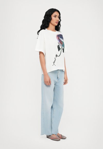 Hvit overdimensjonert t-skjorte med et fargerikt abstrakt design og tekst, parret med lyseblå vide jeans og stripete slip-on sko.