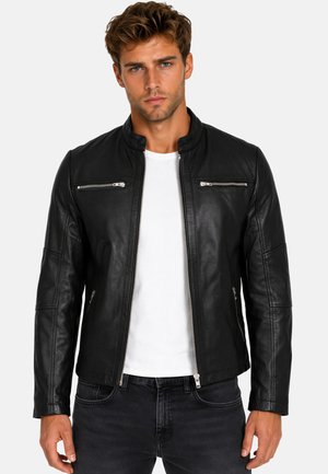 Jonge man met kort krullend haar, draagt een zwart leren jas met ritssluiting over een wit T-shirt en zwarte spijkerbroek, kijkt recht vooruit met een neutrale blik.