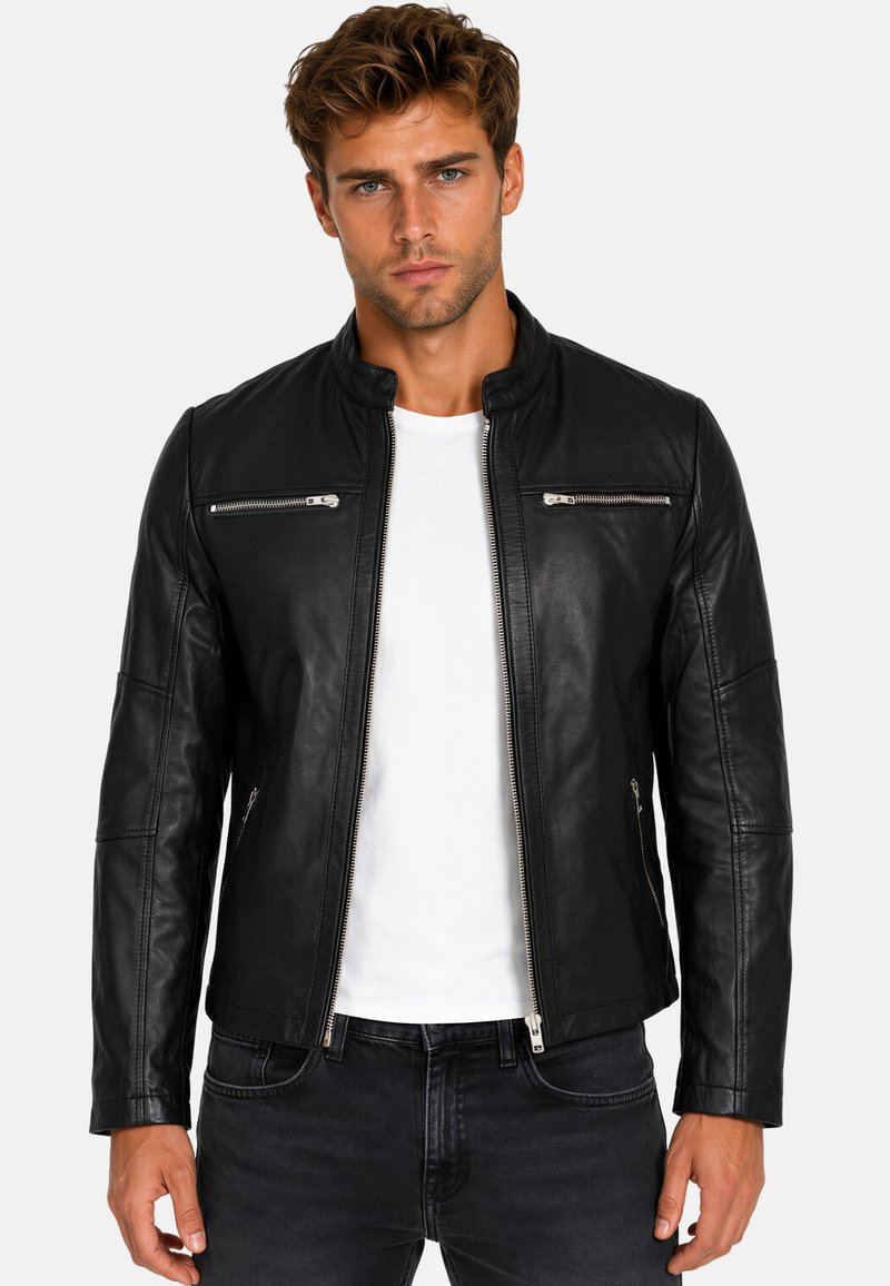 Junger Mann mit kurzen lockigen Haaren, der eine schwarze Lederjacke mit Reißverschluss über einem weißen T-Shirt und schwarzen Jeans trägt, schaut mit neutralem Ausdruck nach vorne.