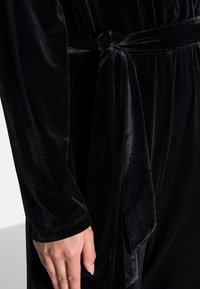 Combinaison en velours noir avec des manches longues, une taille ceinturée et une texture lisse. Le tissu possède un léger éclat et une profondeur de couleur riche.