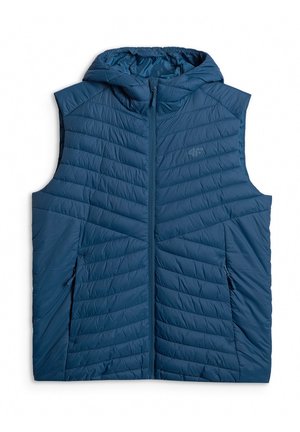 Mężczyzna stoi z rękami w kieszeniach, ubrany w niebieską kamizelkę puchową na szarą bluzę z kapturem i suwakiem oraz dopasowane szare spodnie i białe trampki.