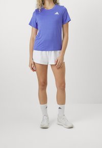 Kvinna i lila Adidas t-shirt, vita shorts, vita Adidas strumpor och vita sneakers stående mot en schlicht vit bakgrund.