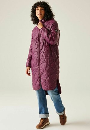 Kobieta ubrana w długą pikowaną fioletową kurtkę z kapturem, obcięte niebieskie dżinsy i brązowe buty trekkingowe, stojąca na jednolitym tle.