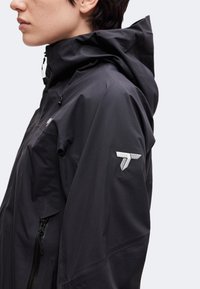 Vue latérale d'une personne portant une veste à capuche noire avec un logo blanc sur le haut du bras et une poche zippée sur la poitrine.