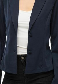 Námořnický modrý blazer s jedním knoflíkem, dlouhými rukávy a měkkou, texturovanou tkaninou. Nosený přes bílý top s černými džíny.