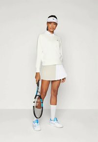 Balts garais piedurkņu krekls, gaiši pelēkas plisētās svārki ar baltiem akcentiem, balta viziera cepure, zila akcentēta tenisa apavi, tur rokās melnu tenisa raketi.