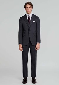 Traje ajustado de color gris oscuro con dos bolsillos frontales, pantalones de corte slim, camisa blanca y una corbata de rayas oscuras. Zapatos de vestir marrones pulidos.