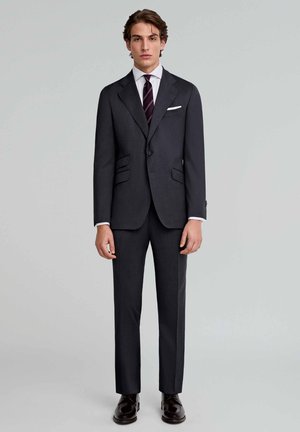 Traje ajustado de color gris oscuro con dos bolsillos frontales, pantalones de corte slim, camisa blanca y una corbata de rayas oscuras. Zapatos de vestir marrones pulidos.