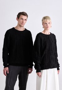 Black knitted sweaters with textured cable patterns, round necklines, and ribbed hems. The male model wears dark pants; the female model wears a white skirt.