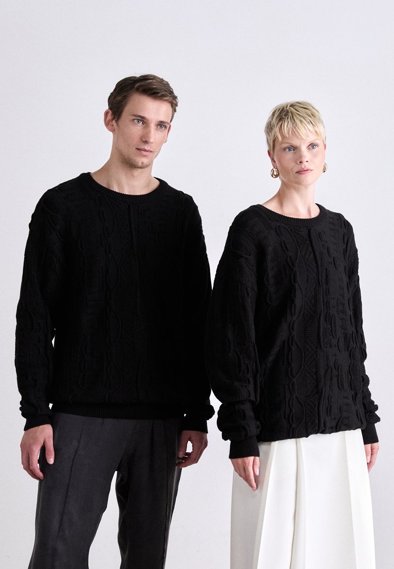 Schwarze Strickpullover mit strukturierten Zopfmustern, runden Ausschnitten und gerippten Säumen. Das männliche Model trägt dunkle Hosen; das weibliche Model trägt einen weißen Rock.