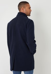 Marineblauer Wollmantel mit hohem Kragen, langen Ärmeln und einer Rückenbelüftung für mehr Bewegungsfreiheit. Sanfte Textur, taillierte Passform.