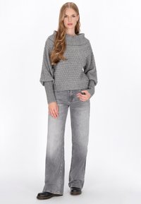 Gråstickad tröja med en bred cowl-hals, texturerad kropp och ribbade ärmar, kombinerad med grå utsvängda jeans och svarta ankelstövlar.
