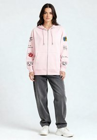 Ružová mikina na zips z mäkkého materiálu, s rôznymi potlačami a vzormi na rukávoch, nosená s voľnými sivými džínsami a bielymi teniskami.