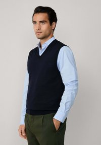 Muž s námorníckym modrým bezrukávovým sveterom s V-výstrihom, oblečený na svetlomodrej dlhé rukávy košeli a olivovozelených nohaviciach, stojí s jednou rukou v vrecku.