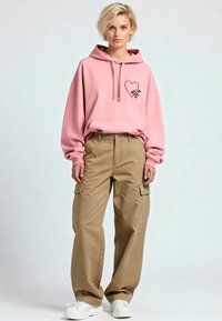 Felpa con cappuccio rosa chiaro decorata con una grafica di un cuore e una spilla di sicurezza, abbinata a pantaloni cargo beige larghi. Scarpe da ginnastica bianche completano l'outfit.