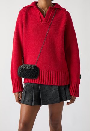 Vrouw draagt een felrode gebreide trui met V-hals, een zwarte geplooide minirok en een kleine zwarte crossbodytas met een kettingriem.