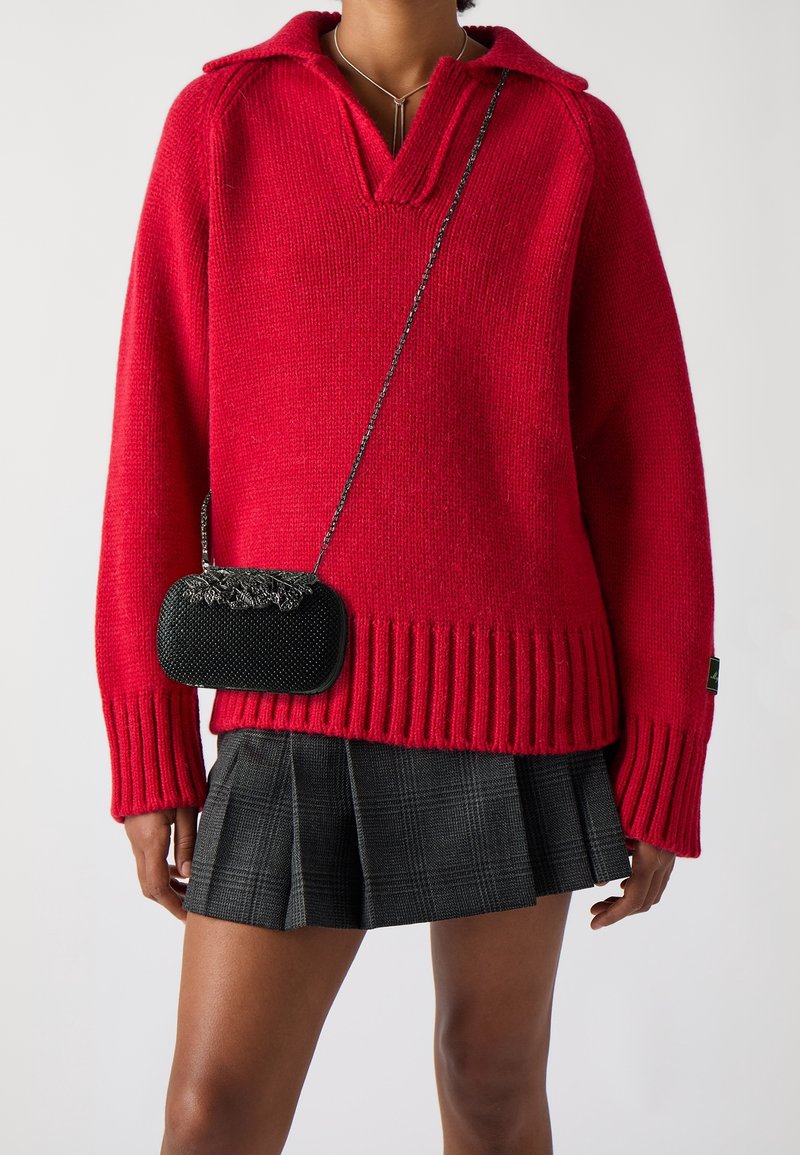 Femme portant un pull en maille rouge vif à col en V, une mini-jupe plissée noire et un petit sac bandoulière noir avec une bandoulière chaîne.