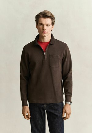 Jonge man die een donkerbruine pullover met kwart-ritssluiting draagt over een rood overhemd en donkere spijkerbroek, staand tegen een effen lichte achtergrond.