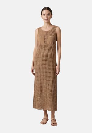 Donna con capelli scuri raccolti in uno chignon, indossa un lungo abito marrone senza maniche in maglia con due tasche sul petto e sandali beige, in piedi su sfondo bianco.
