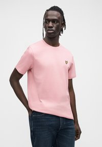 Lyhythihainen vaaleanpunainen puuvillainen T-paita, jossa on pyöreä pääntie ja pieni keltainen embleemi vasemmassa rinnassa. Yhdistetty tummiin farkkuihin.