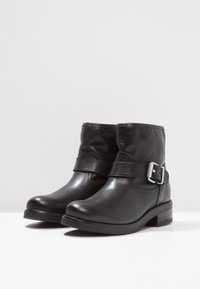 Botines de cuero negro hasta el tobillo con punta redondeada, detalle de correa y acentos de hebilla plateada. Parte superior texturizada y diseño de tacón bajo y cuadrado.