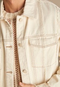 Chaqueta de denim beige con costuras marrones en contraste, dos bolsillos frontales y textura suave. Debajo, un top beige de punto con un patrón de malla.