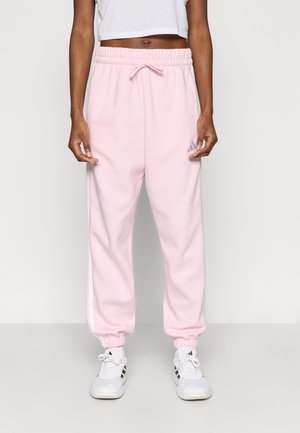 Person iført lyserøde sweatpants med hvide sidelister, hvide sneakers og en hvid croptop mod en ensfarvet baggrund.