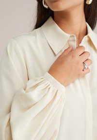 Vrouw in een ivoorwitte blouse met pofmouwen, die de kraag met één hand vasthoudt en een zilveren ring en een gemarmerde oorbel toont.
