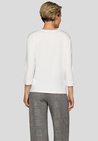 Person mit kurzen Haaren, der ein einfaches weißes Langarmshirt und graue karierte Hose trägt, mit dem Rücken zum Betrachter vor einem schlichten Hintergrund.