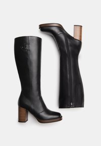 Bottes hautes en cuir noir avec talons blocs empilés marron, fermeture éclair latérale, et logo subtil près du sommet, présentées sur un fond blanc.