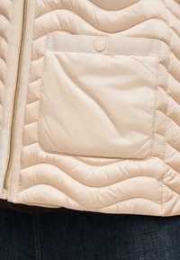 Τμήμα μπεζ μπουφάν με καπιτονέ σχέδιο, εμπρός φερμουάρ και πλαϊνή τσέπη με καπάκι ασφαλισμένο με στρογγυλό κουμπί, φορεμένο πάνω από σκοτεινό τζιν.