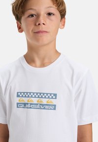 Weißes Baumwoll-T-Shirt mit einem grafischen Druck, der blaue, gelbe und graue Elemente umfasst, darunter ein kariertes Muster und das 'Quiksilver'-Logo.