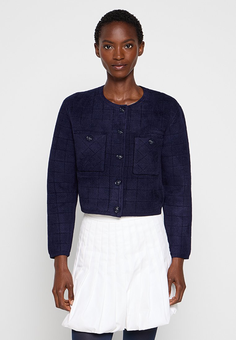 Granatowa pikowana kurtka o cropped kształcie, z okrągłym dekoltem, przednim zapięciem na guziki, dwoma kieszeniami na piersiach oraz ściągaczami na mankietach. Miękka, teksturowana tkanina.