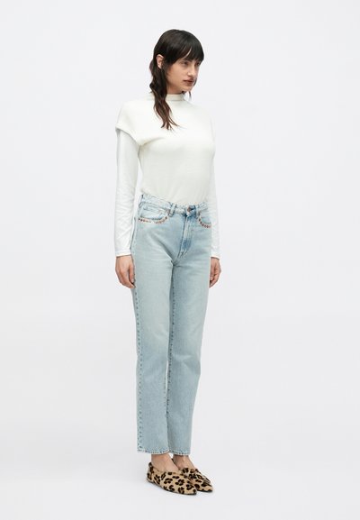 Λευκή υφασμάτινη μπλούζα με ψεύτικο λαιμό, τζιν παντελόνι ελαφριάς πλύσης με ίσια γραμμή και διακοσμητικές ραφές, και μοκασίνια με λεοπάρ σχέδιο.