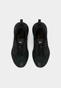 Ζεύγος μαύρων αθλητικών παπουτσιών Lacoste με χρυσλό λογότυπο κροκοδείλου, σχέδιο με κορδόνια και θηλιές έλξης σε λευκό φόντο.