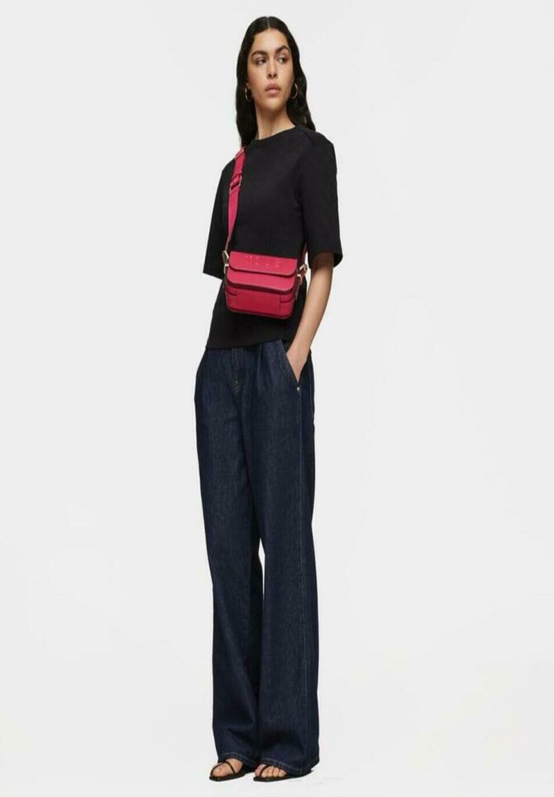 Bolso cruzado rojo con un acabado texturizado, llevado sobre una blusa negra y pantalones anchos de mezclilla azul, complementado con sandalias negras.