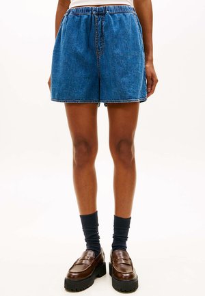 Blaue Denim-Shorts mittlerer Länge, kombiniert mit schwarzen Socken und braunen klobigen Loafers an einer stehenden Person vor einem einfarbigen Hintergrund.