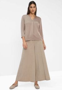 Giovane donna in piedi che indossa una blusa taupe con scollo a V, pantaloni beige a gamba larga e scarpe beige, su uno sfondo neutro.