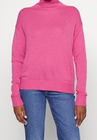Roze coltrui van ribgebreid katoen, met lange mouwen en brede manchetten, en een klein merkdetail op de borst.