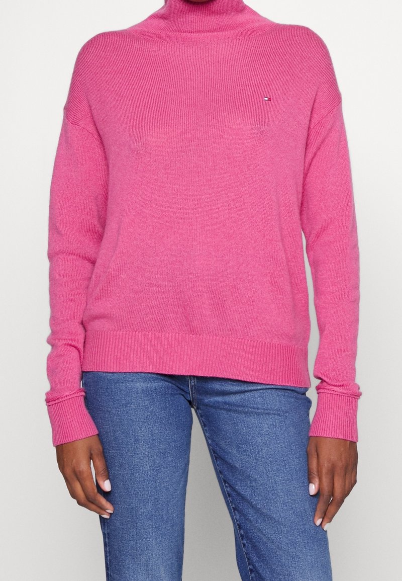 Roze coltrui van ribgebreid katoen, met lange mouwen en brede manchetten, en een klein merkdetail op de borst.