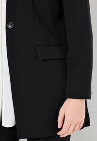 Cappotto lungo nero con un design elegante, caratterizzato da una chiusura a un bottone, tasche laterali e una texture liscia. Mostrato con una camicia interna bianca.