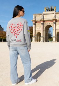 Femme aux longs cheveux noirs portant des lunettes de soleil, un sweatshirt gris avec des cœurs rouges et le texte "Sweet heart", debout près d'une arche en pierre ornée en plein jour.