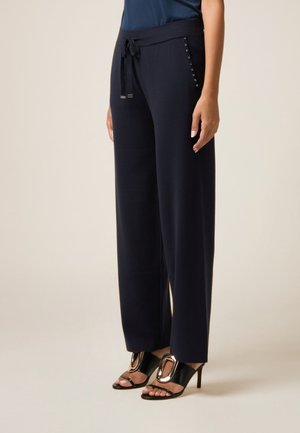 Marineblaue weit geschnittene Hose mit einem Kordelzugbund und dekorativen metallischen Akzenten an den Taschen, kombiniert mit schwarzen, hohen Sandalen. Glatte Textur.
