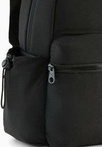 Sac à dos noir en tissu lisse, avec des zips proéminents et un cordon bungee sur la poche latérale pour un rangement supplémentaire.