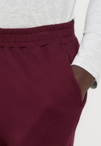 Κοντινό πλάνο ενός ατόμου που φοράει παντελόνι με ελαστική μέση μπορντό, με το δεξί χέρι στην τσέπη και ένα ανοιχτό γκρι μακρυμάνικο μπλουζάκι.