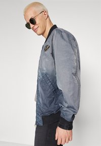 Veste bomber bleue avec une texture lisse, poignets et ourlet côtelés, arborant un patch de marque sur la poitrine. Portée avec des lunettes de soleil rondes.